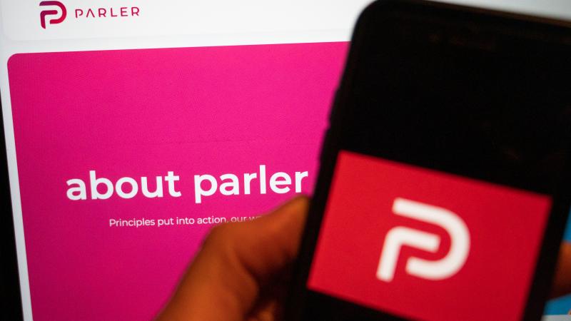 On a mobile phone, the logo of the social media platform Parler is displayed. In the background on a screen is the platform's website.