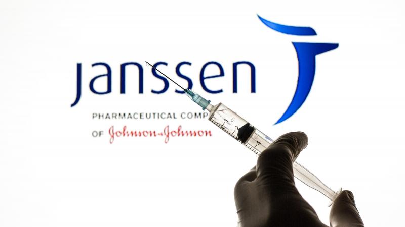 In this photo illustration, a close up of a hand holding a medical syringe in front of the Janssen vaccine logo.