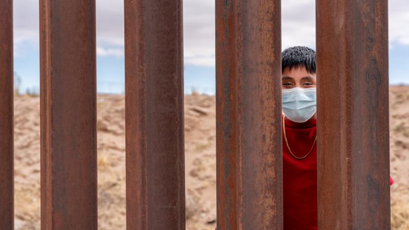 Migrant looks through border wall divide while wearing COVID mask