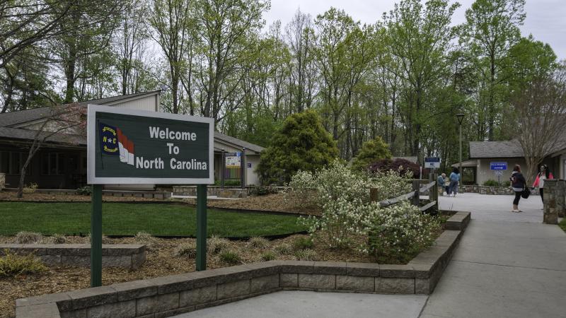 A Welcome to North Carolina sign at a rest area with travelers relaxing in the background.
